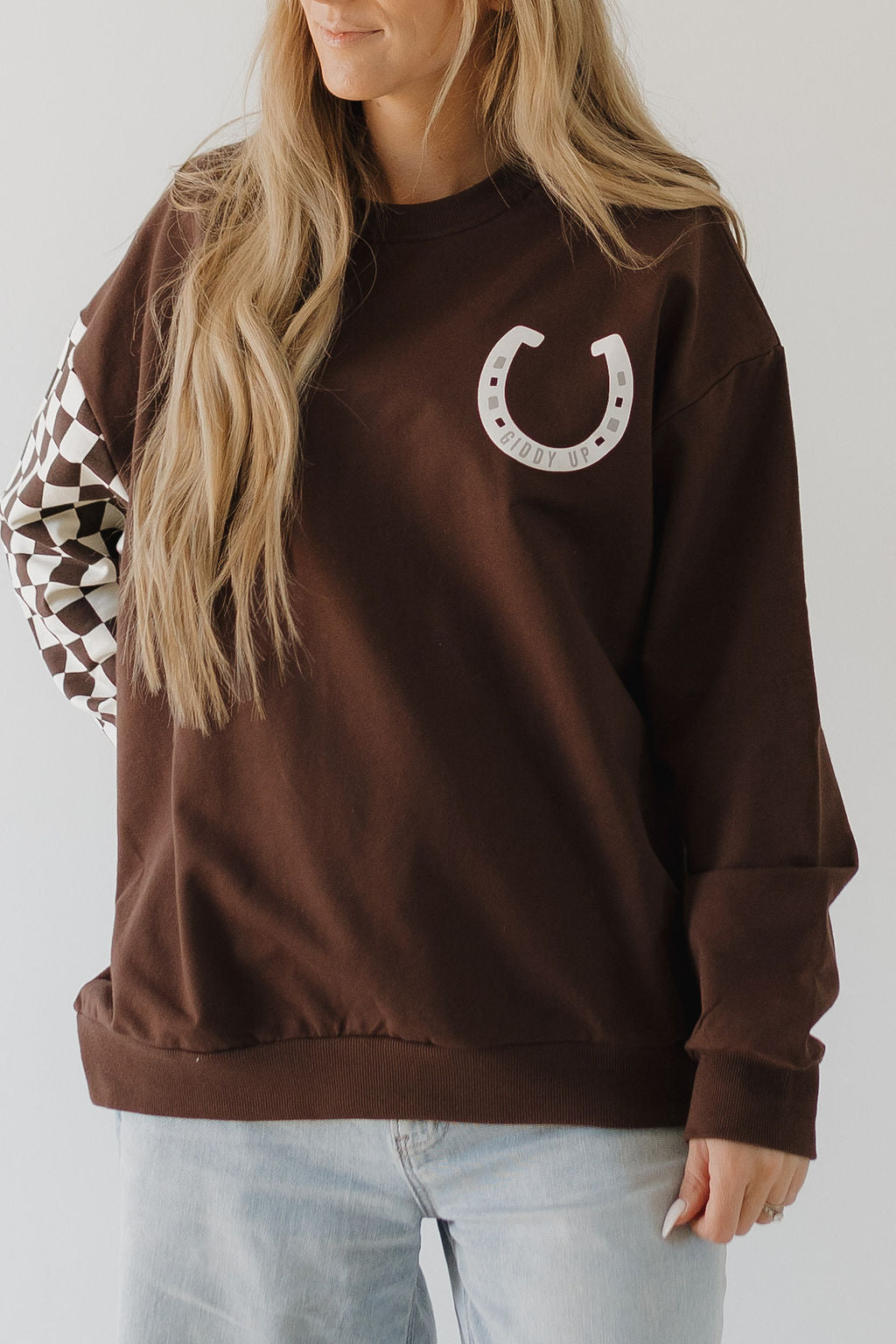 A person with long hair wears the "Adult Sweatshirt | Giddy Up" from forever french baby, showcasing a horseshoe graphic and the words "Giddy Up," with one sleeve in a black and white checkered pattern, paired with light-colored jeans.