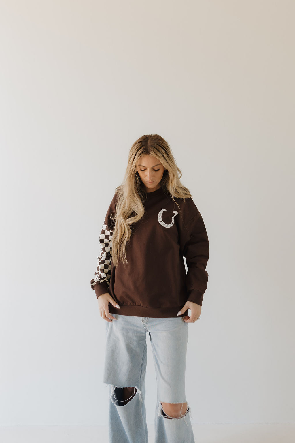 A woman with long blonde hair models a brown Giddy Up sweatshirt by forever french baby, showcasing a horseshoe design and checkered sleeves. Paired beautifully with light blue ripped jeans, she stands against a plain white backdrop, gazing downward to capture casual style harmony.