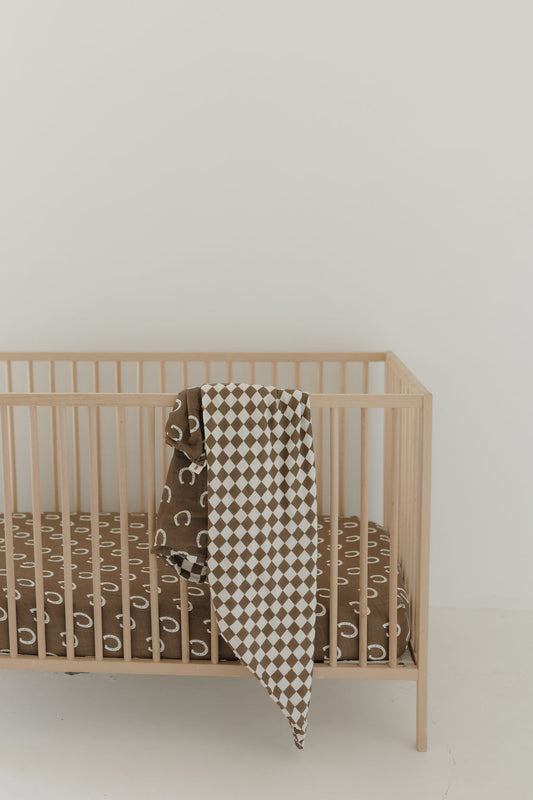 A minimalist wooden crib holds a brown abstract patterned mattress and is adorned with the Muslin Quilt | Giddy Up/Dutton from forever french baby, showcasing a brown and white checkered design. The backdrop of an off-white wall creates a serene nursery setting.