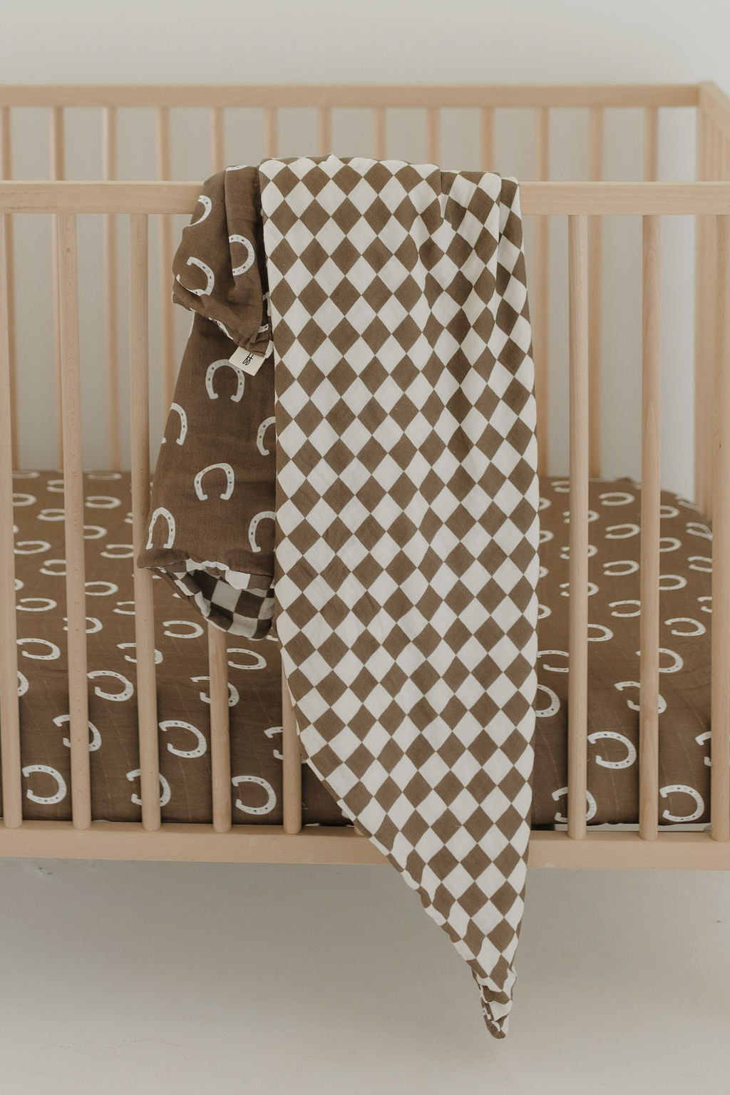 In a cozy nursery, the wooden baby crib showcases the forever french baby Muslin Quilt in Giddy Up/Dutton style, featuring a brown and white checkered design. The mattress has a brown sheet with charming white horseshoe patterns against a plain white background.