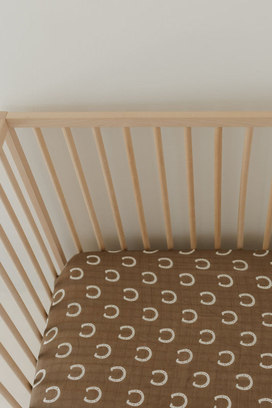 A wooden baby crib with a brown mattress adorned in white horseshoes gets a cozy upgrade with the "Giddy Up" Muslin Crib Sheet from forever french baby, adding charm to the nursery against a light-colored wall.