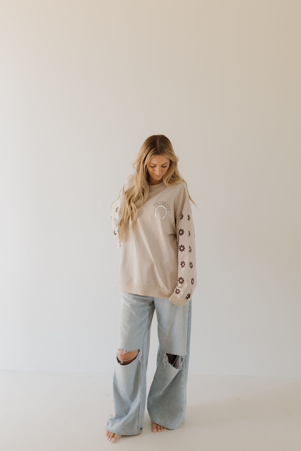 A woman wearing a forever french baby Adult Sweatshirt from the Howdy Collection, with floral sleeves, stands barefoot on a plain backdrop in light blue ripped jeans, capturing a coordinated moment as she looks down.