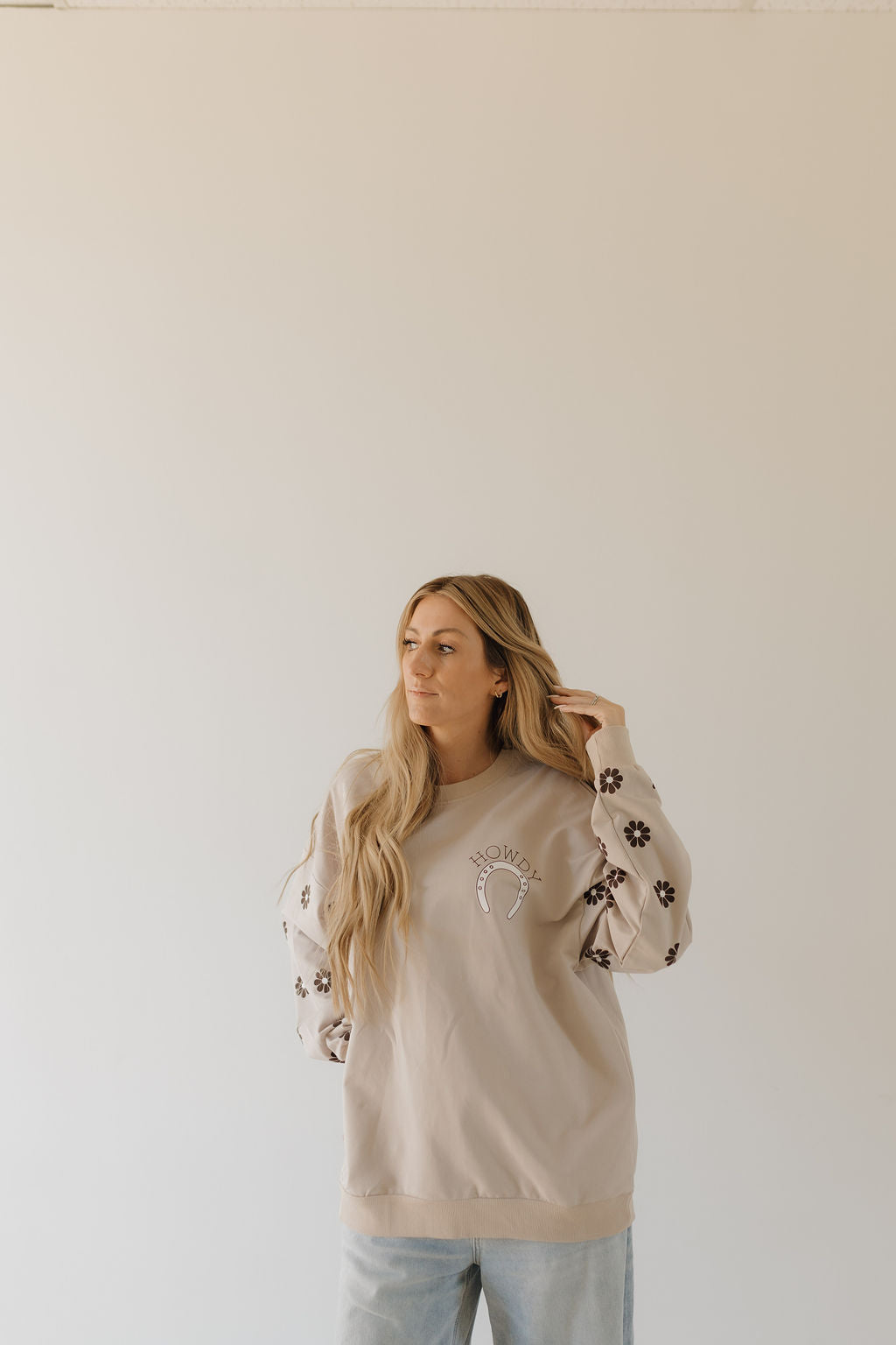 Against a plain background, a person with long light brown hair, looking to the side and touching their hair, wears a cozy Adult Sweatshirt from forever french baby’s Howdy Collection, featuring a crescent moon design and floral sleeve patterns.