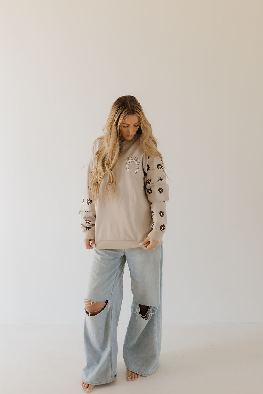 A woman with long hair stands barefoot in a cozy forever french baby sweatshirt from the Howdy Collection, featuring floral-patterned sleeves. Wearing light blue ripped jeans, she gazes downward against a plain white background.