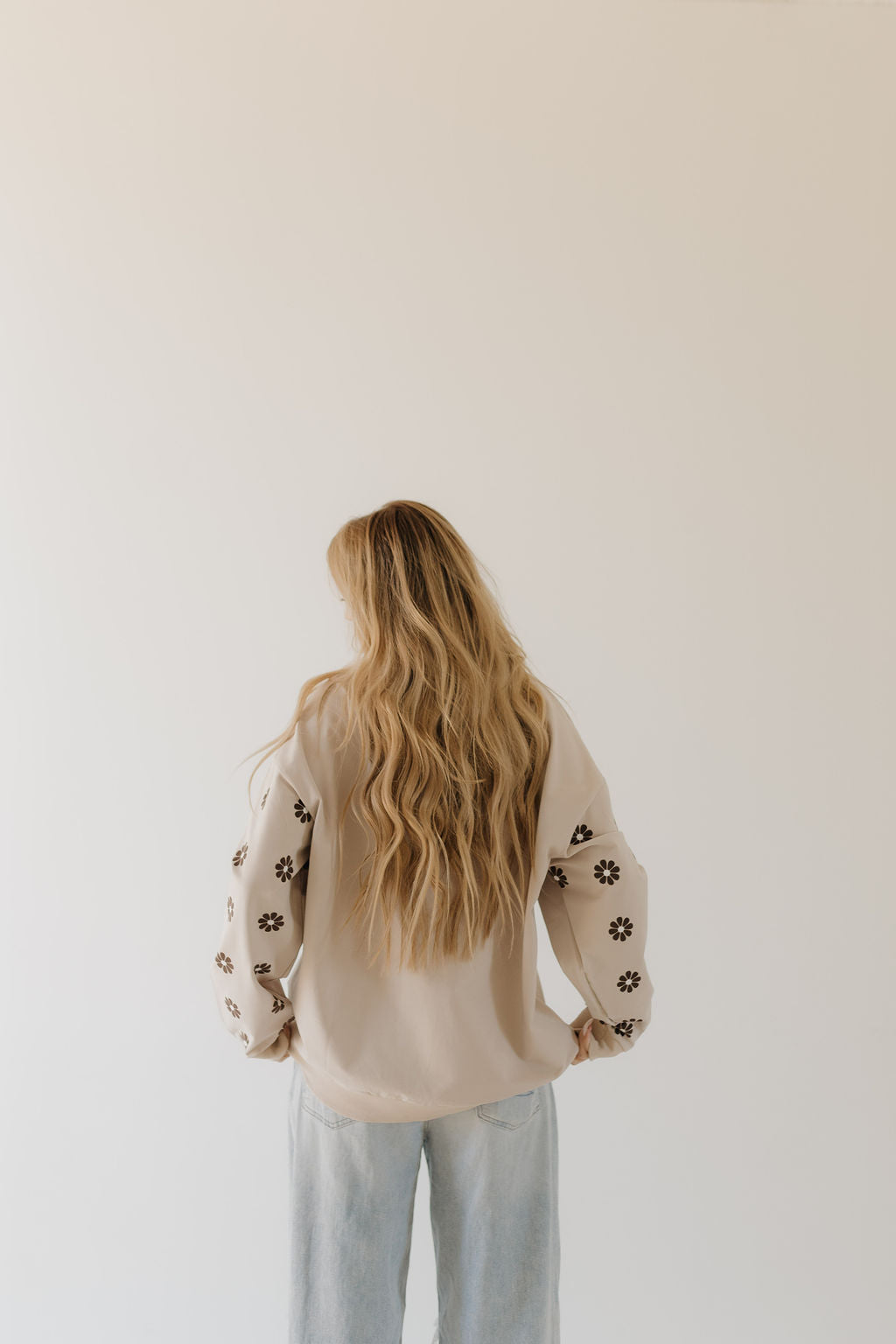 A person with long, wavy blond hair faces away wearing the Adult Sweatshirt from forever french baby's Howdy Collection, featuring floral sleeve patterns, paired with light blue jeans. The plain, light-colored wall background adds a touch of subtle elegance.