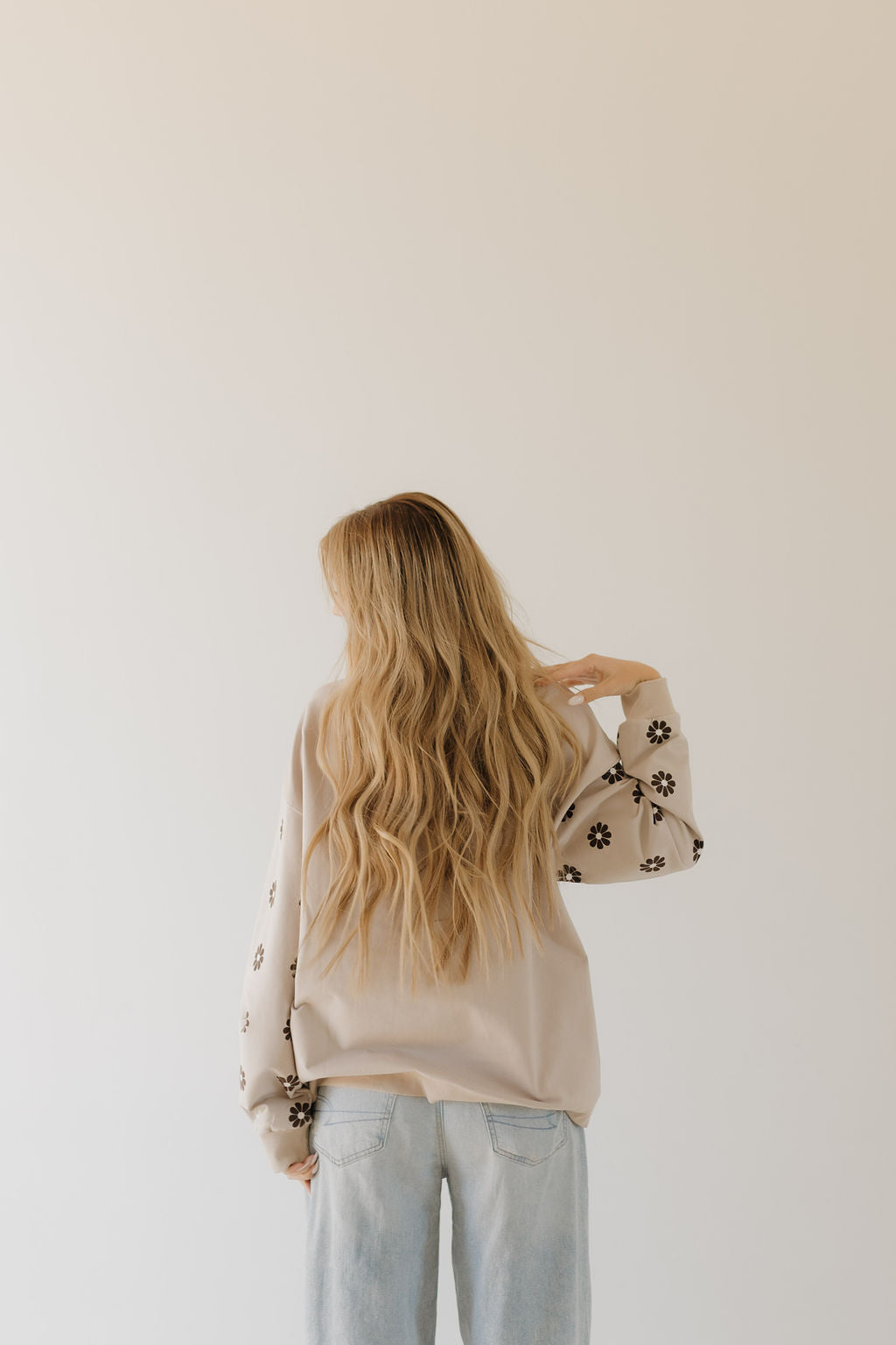 A woman with long, wavy blonde hair wears a cozy Adult Sweatshirt from the Howdy collection by forever french baby, featuring floral sleeve patterns. She pairs it with light blue jeans and faces away from the camera against a neutral backdrop.