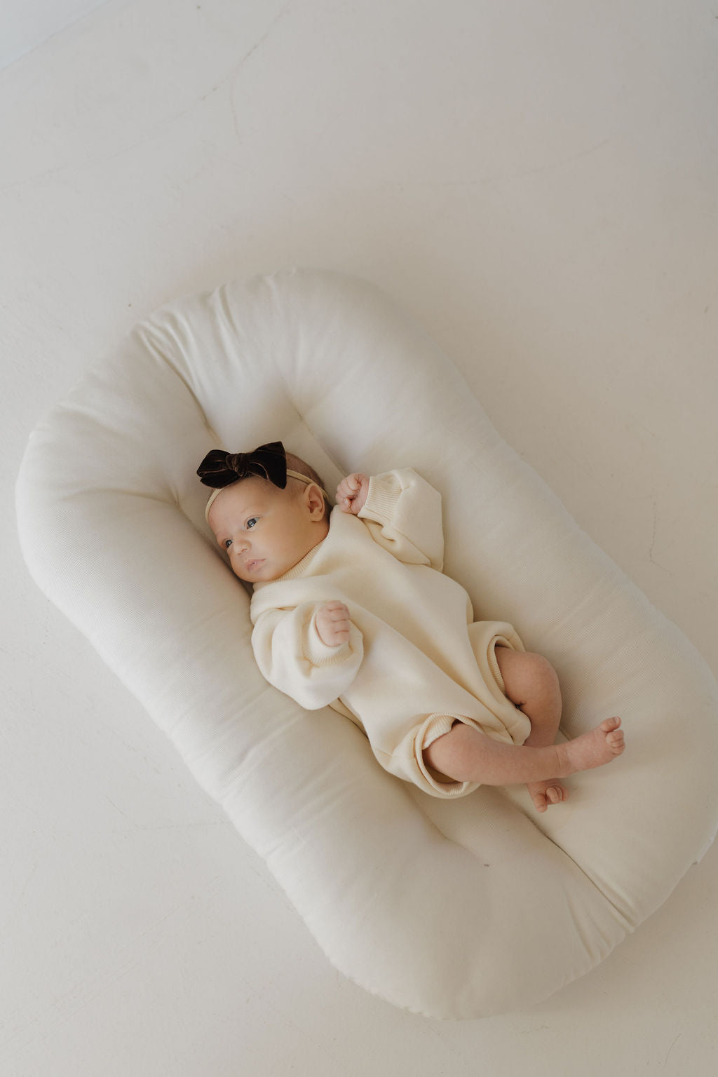 A baby in a cream-colored outfit and black bow headband lies on a white cushioned lounger. The Sweatshirt Romper | Oat by forever french baby ensures easy diaper changes, with tiny hands and feet enhancing the cuteness against the smooth, light background.