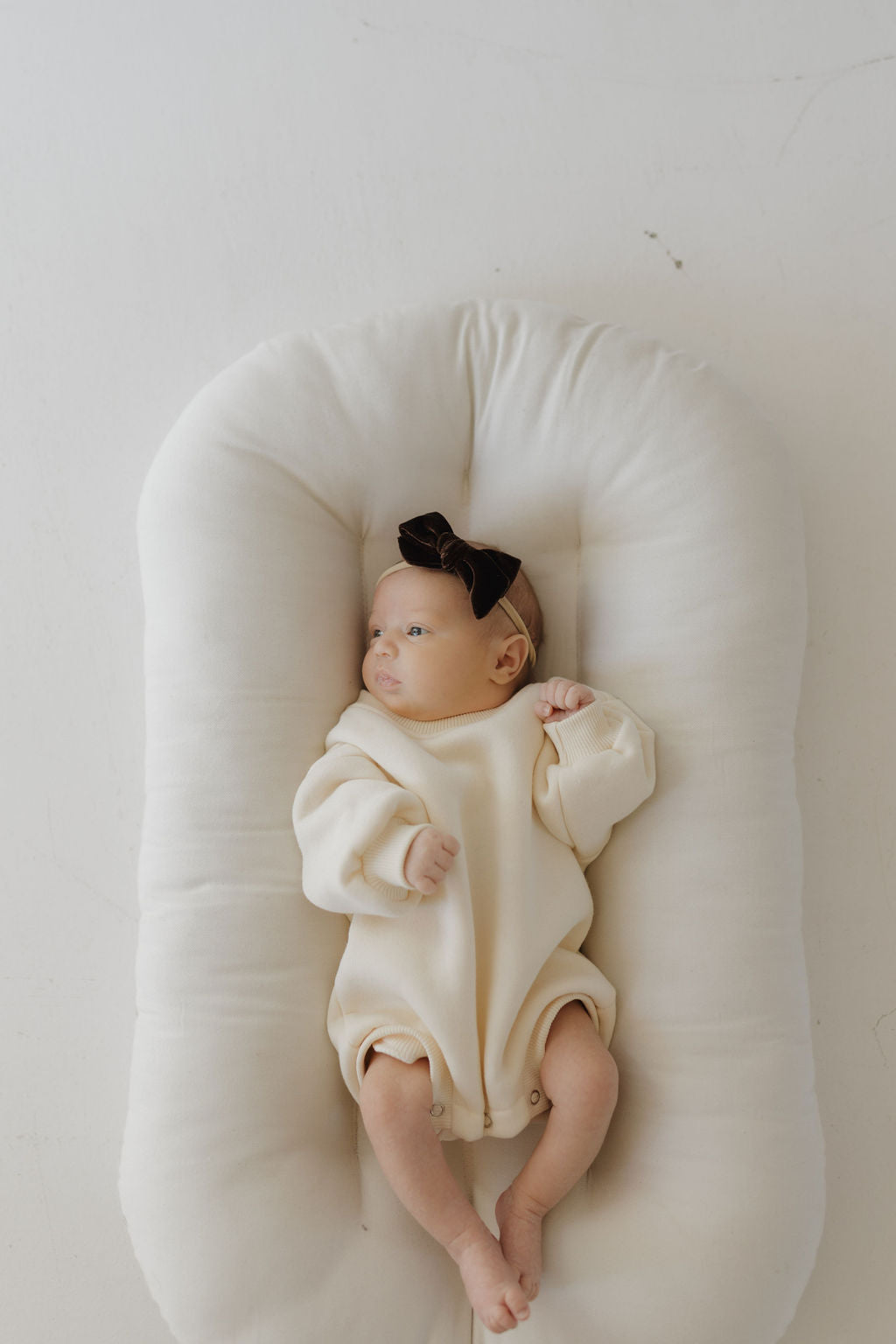 A baby in a forever french baby Sweatshirt Romper | Oat, paired with a dark bow, rests on a soft oval cushion. A plain white backdrop adds serenity as the baby gazes upward and raises one hand, ideal for easy diaper changes.