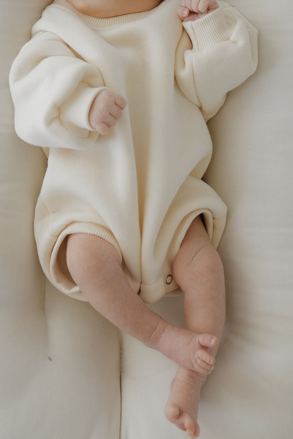 A baby rests comfortably in a forever french baby Sweatshirt Romper | Oat, with partially visible hands and one hand slightly curled. The cozy, oversized design ensures easy diaper changes as its legs cross at the ankles in a relaxed pose.