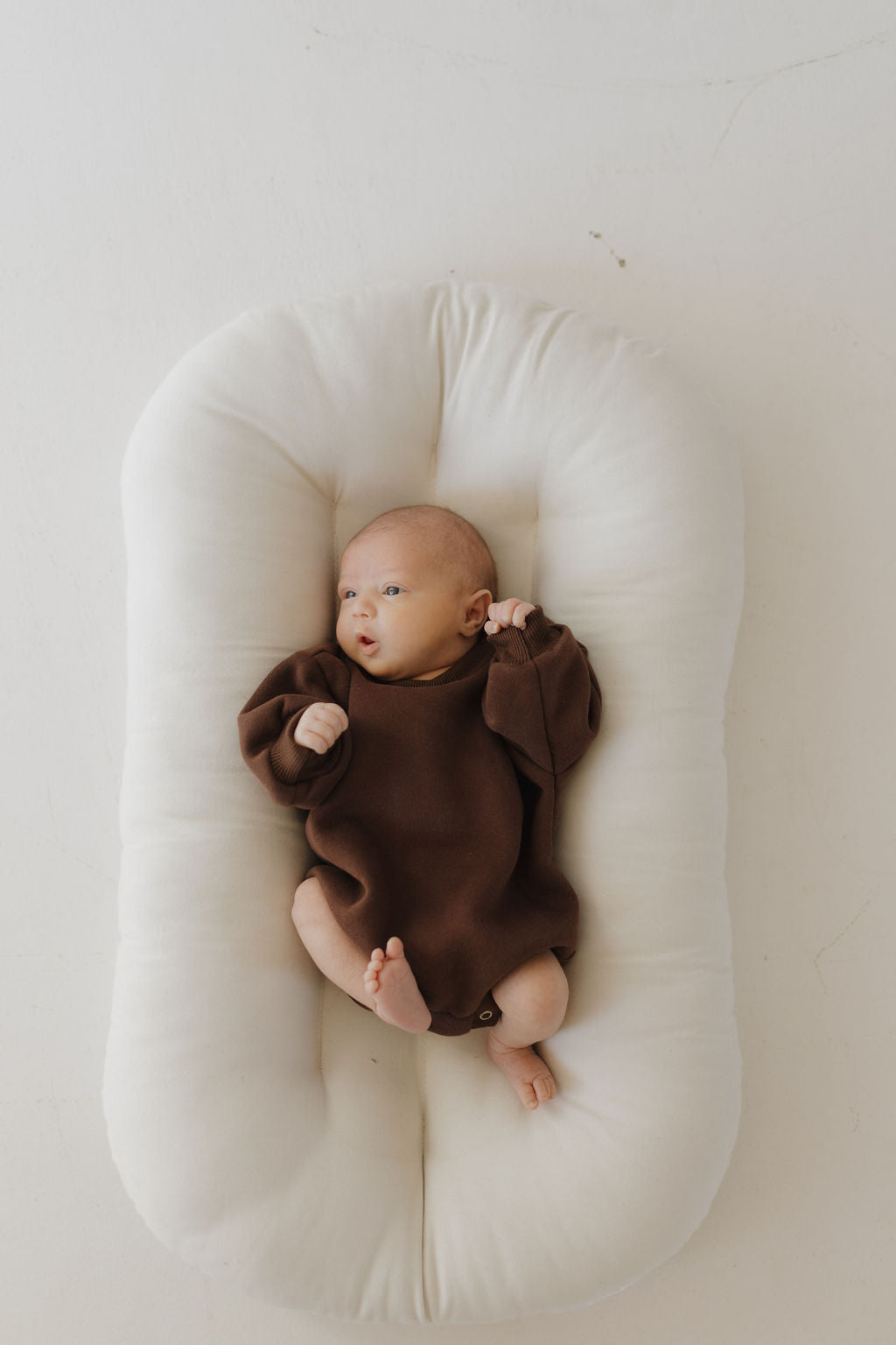 A baby in a neutral-toned "Sweatshirt Romper | Driftwood" from forever french baby rests in a soft, white nest. The cozy romper allows easy diaper changes as the infant looks up with raised hands against a minimalist light backdrop.