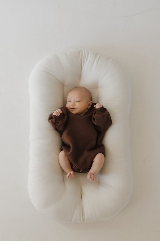 A newborn lies on a soft white cushion, wearing the forever french baby Sweatshirt Romper in Driftwood, ideal for easy diaper changes. The baby's arms are slightly raised in contentment against a plain, pale background.