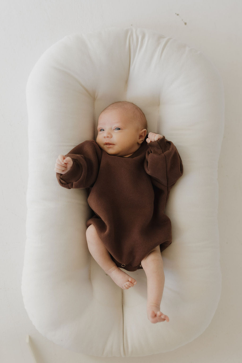 A baby in a brown Sweatshirt Romper | Driftwood from forever french baby lies on a soft white cushion, looking relaxed with one arm raised and small bare feet visible. This neutral-toned romper is designed for easy diaper changes and blends perfectly with the calm background.