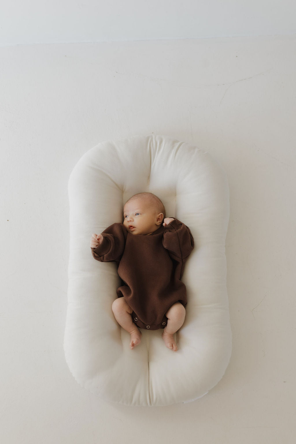 A baby in a Driftwood Sweatshirt Romper by forever french baby lies on a soft, cream cushion. The outfit eases diaper changes. Slightly off-center with one arm raised, the scene is minimalistic against a neutral background.