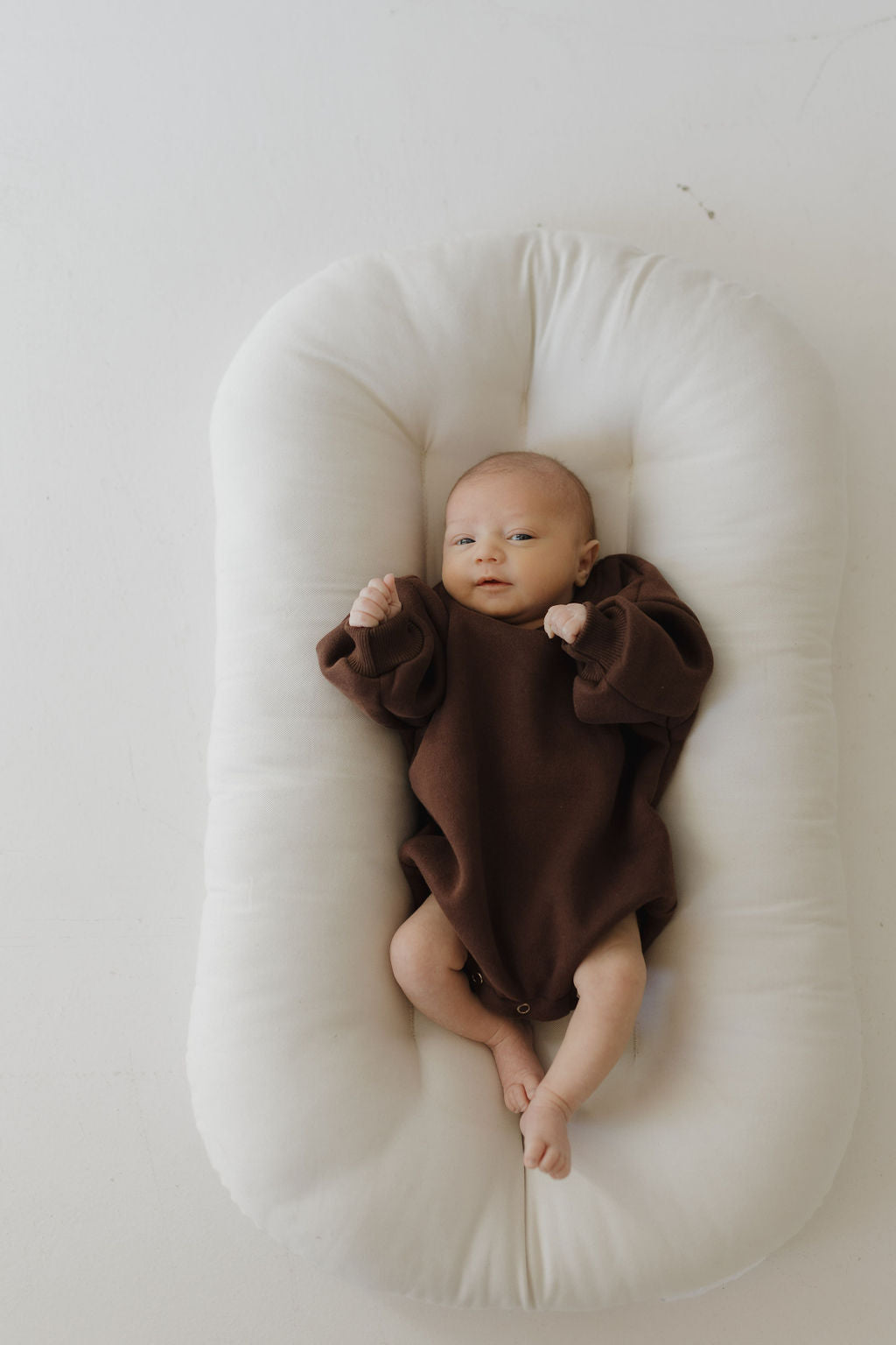 A baby in a Sweatshirt Romper in Driftwood from forever french baby lies on a soft, white cushion. The baby's fists are slightly raised with a calm gaze upward, set against a light background that enhances the peaceful atmosphere perfect for easy diaper changes.