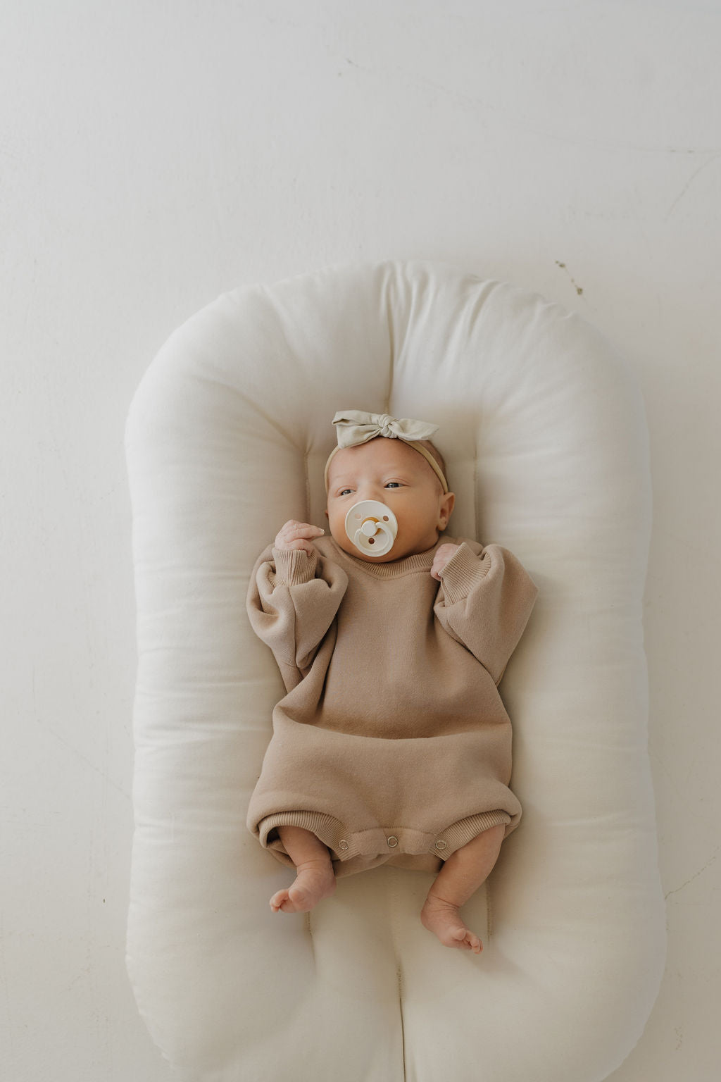 A newborn rests on a soft, white cushion, clad in a forever french baby Sweatshirt Romper in Fawn with a matching headband, holding a pacifier. The romper is designed for convenient diaper changes. The backdrop is a plain light hue.