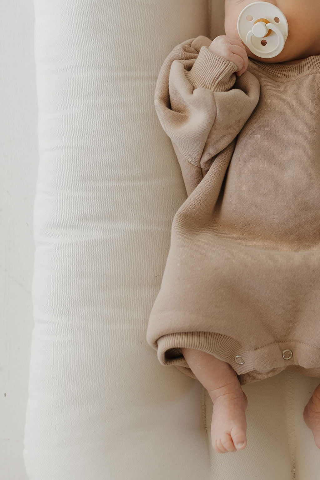 A newborn to 24-month-old baby lies on a white cushion, sucking a pacifier, cozy in forever french baby's neutral-toned Sweatshirt Romper | Fawn. This outfit has oversized sleeves and is designed for easy diaper changes, enhancing the serene ambiance.