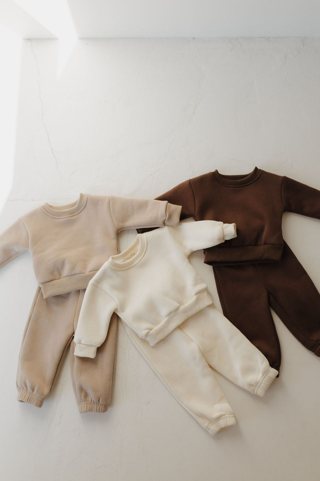 Three sets of forever french baby Sweat Set | Fawn children's clothing are displayed on a light surface. Each set, in beige, cream, and brown, includes a long-sleeve top and pants made from super soft material with an elastic waistband for cozy casual wear.