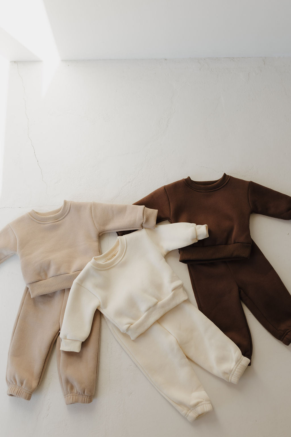 Three toddler-sized sweat sets from forever french baby's "Sweat Set | Oat" collection are displayed on a light surface. These sets, in shades of beige, cream, and dark brown with elastic waistbands, highlight their super soft material for cozy casual wear.