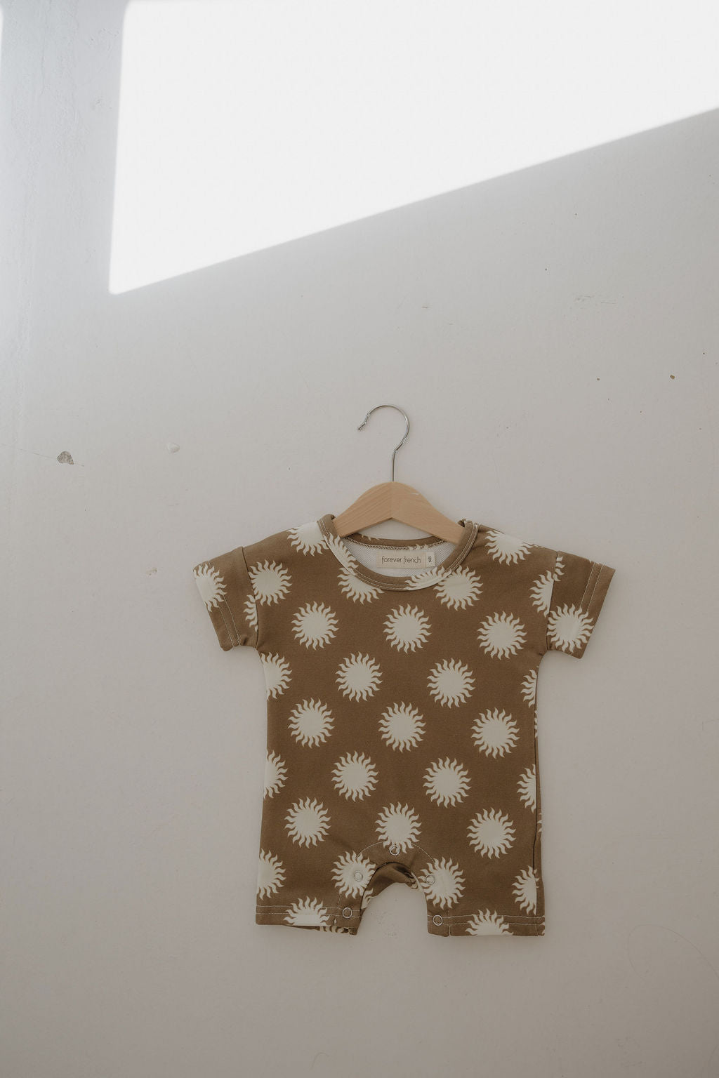 The forever french baby French Terry Romper | Sol, a breathable brown short-sleeve romper with a white sun pattern, hangs on a wooden hanger against a plain white wall bathed in soft natural light.