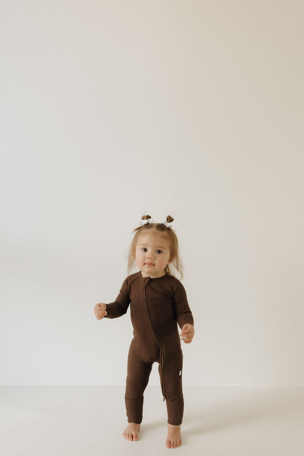 A toddler in a brown, hypo-allergenic "Bamboo Zip Pajamas | Coffee Bean" from forever french baby stands against a plain background. The child has small pigtails and gazes curiously at the camera.