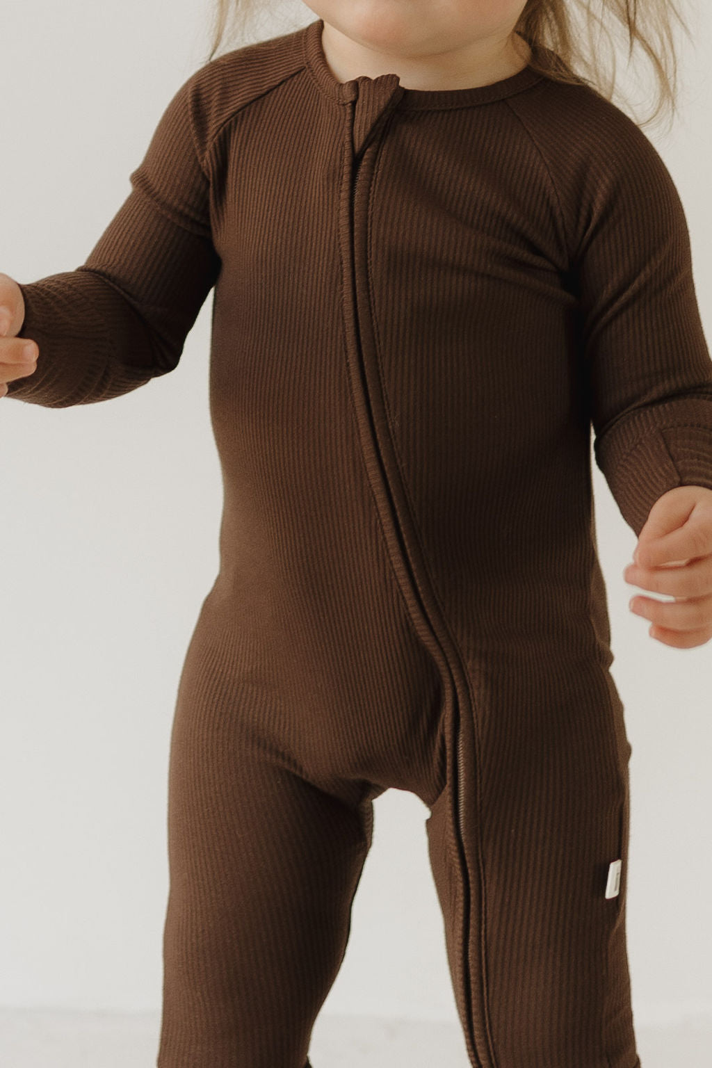 A child wearing forever french baby's Bamboo Zip Pajamas in Coffee Bean stands against a light background, showcasing the breathable ribbed one-piece with a front zipper, their hands gesturing slightly.