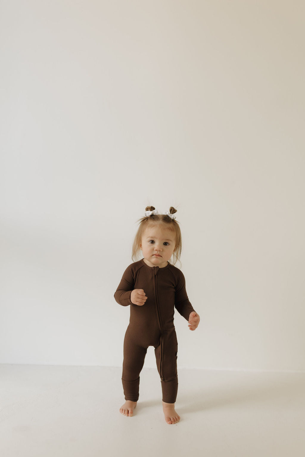 A toddler with two small buns stands barefoot on a light floor, wearing forever french baby's Bamboo Zip Pajamas in Coffee Bean. She looks straight ahead against a plain, light-colored background.