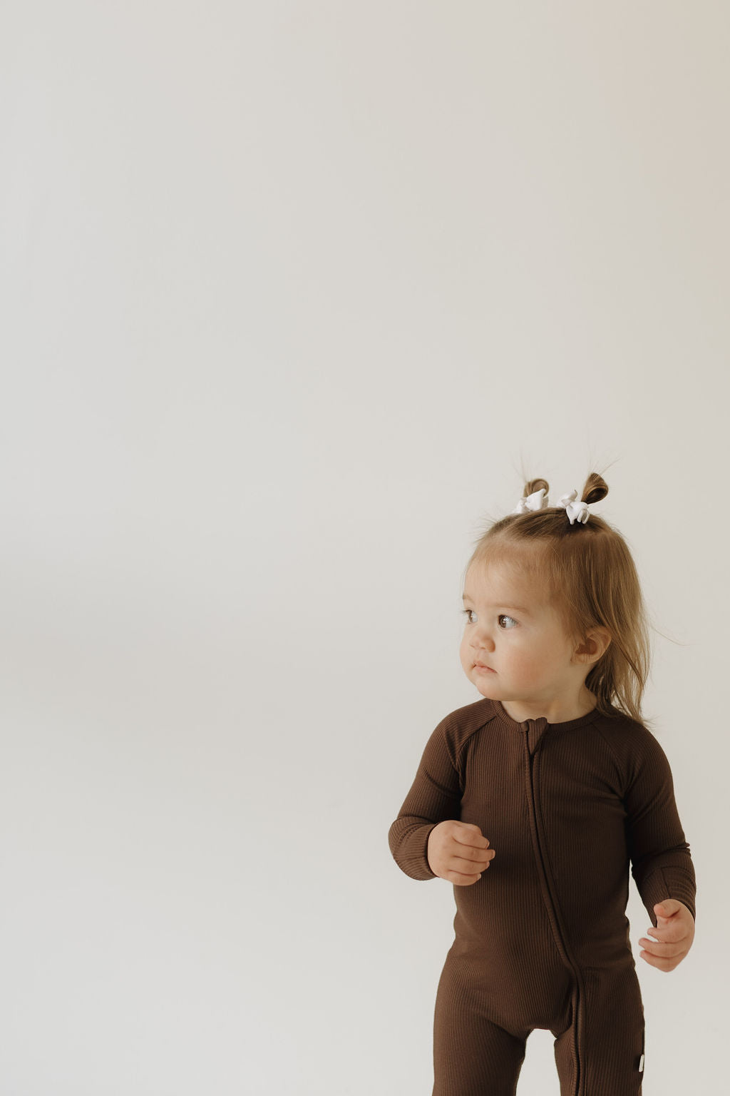 A child in forever french baby Bamboo Zip Pajamas | Coffee Bean stands against a white background. Their hair is styled in two buns with white bows as they gaze to the side curiously.