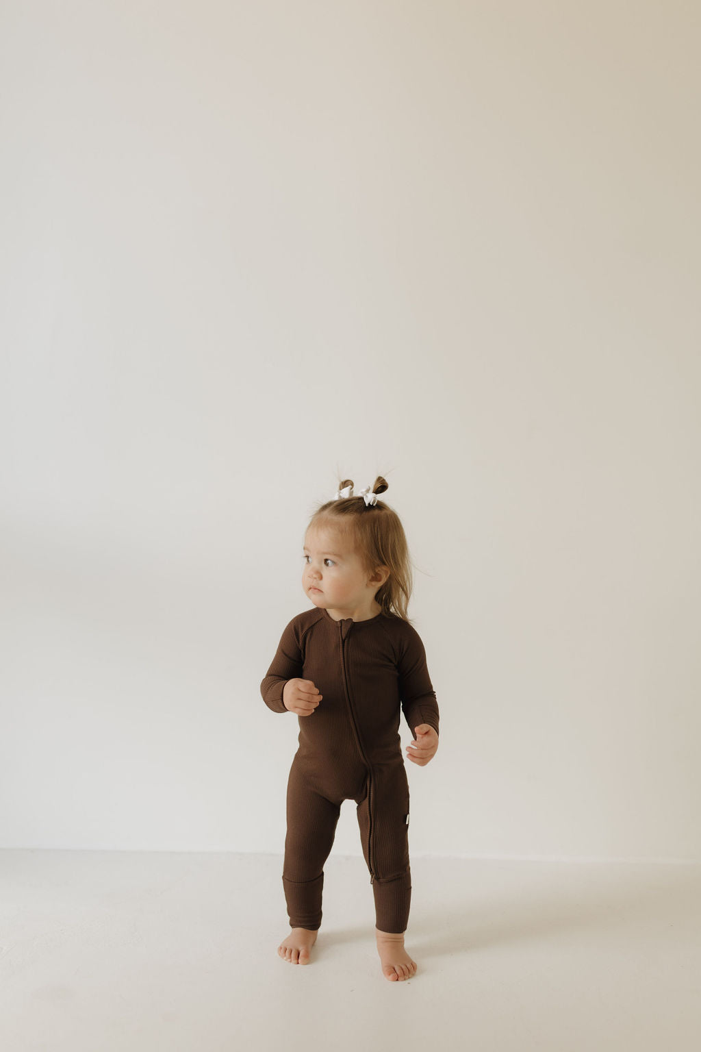 A toddler in forever french baby's Coffee Bean bamboo zip pajamas stands barefoot on a white floor with a plain background. Sporting two small pigtails tied with white bows, the child gazes to the side. .