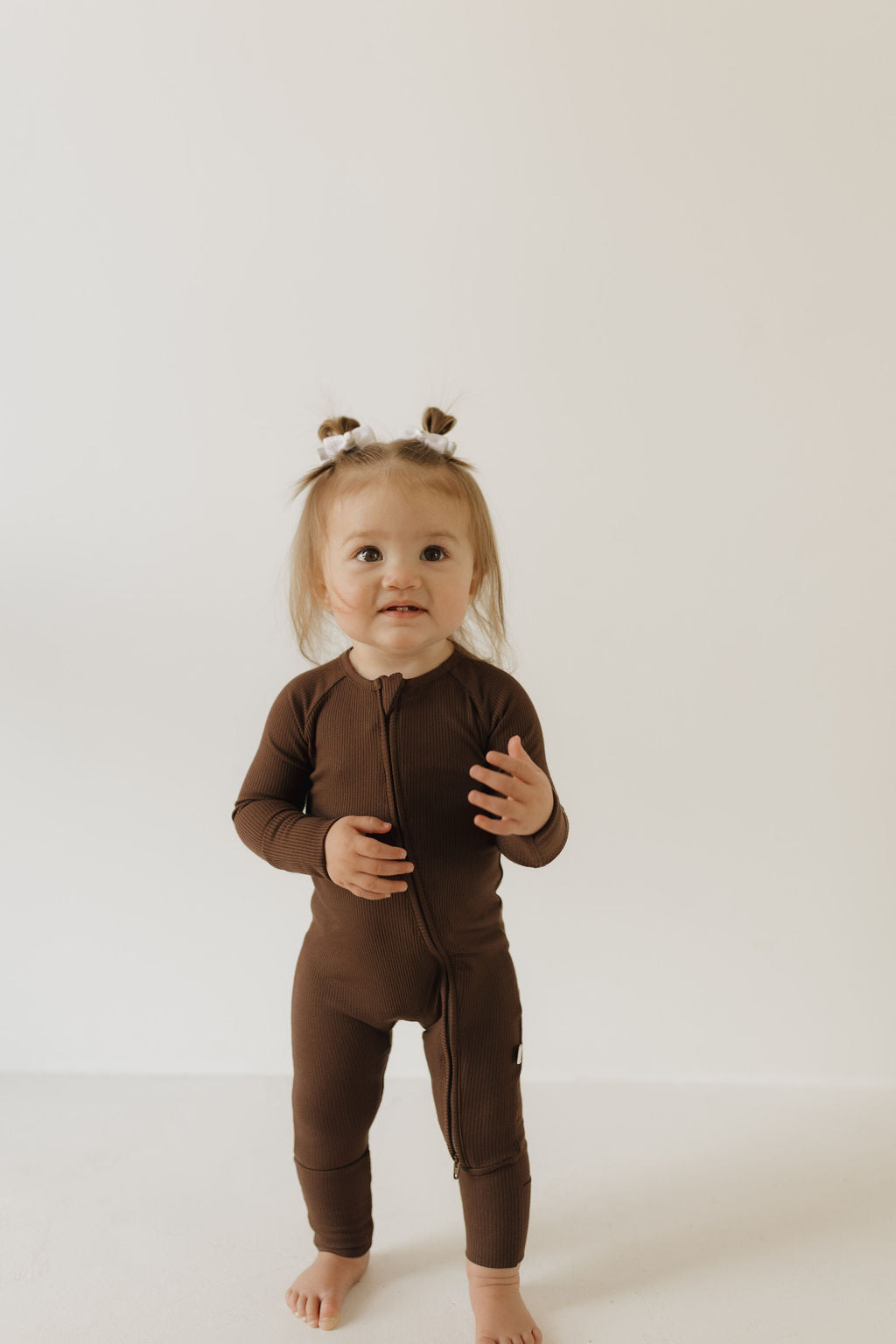 A toddler with two small hair buns stands against a plain white backdrop, barefoot in forever french baby's Coffee Bean Bamboo Zip Pajamas, featuring brown long sleeves and made of breathable fabric, as they face the camera with a neutral expression.