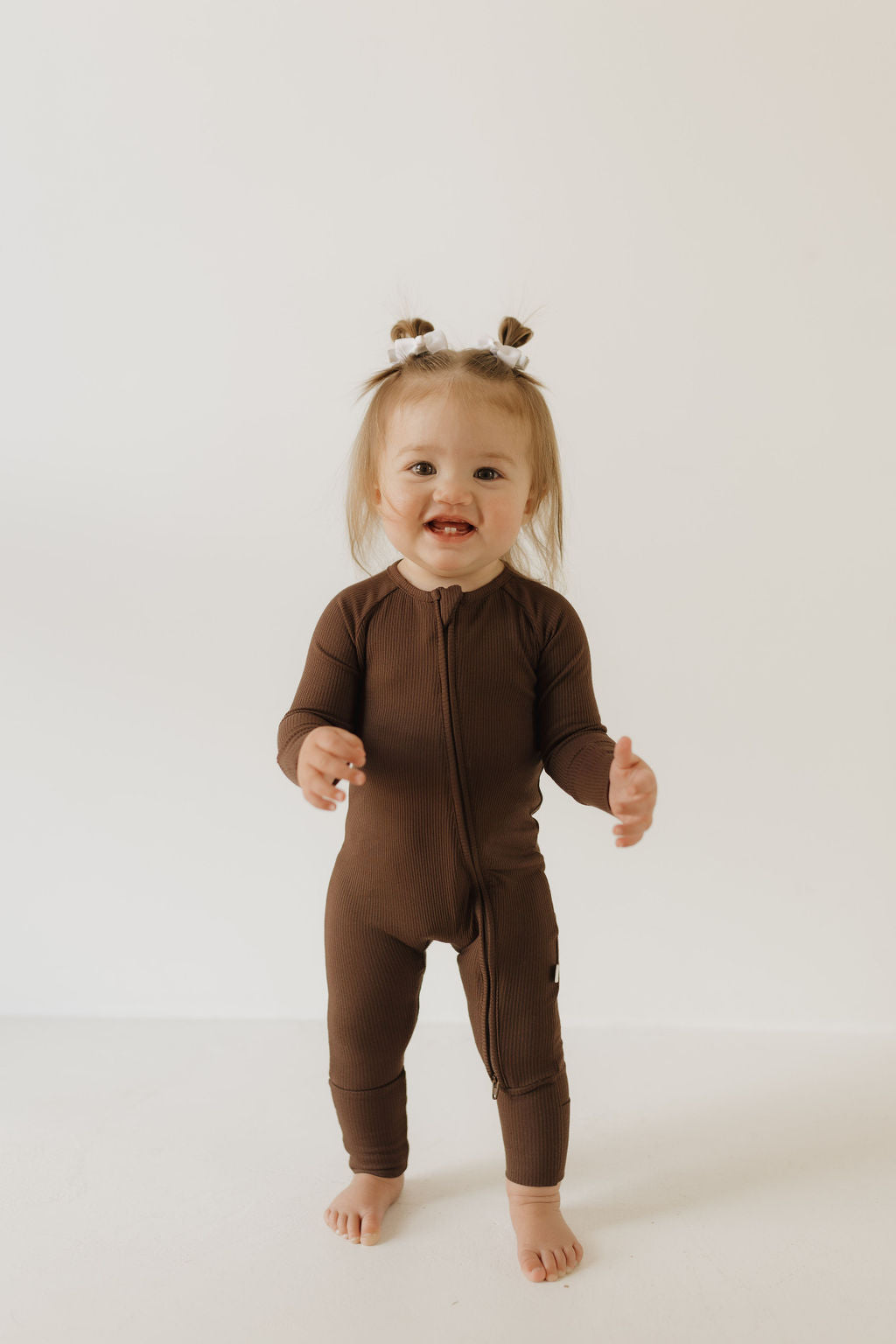 A toddler in a forever french baby Bamboo Zip Pajama in Coffee Bean stands on a white floor, with two small buns in their hair, smiling against a plain white background.