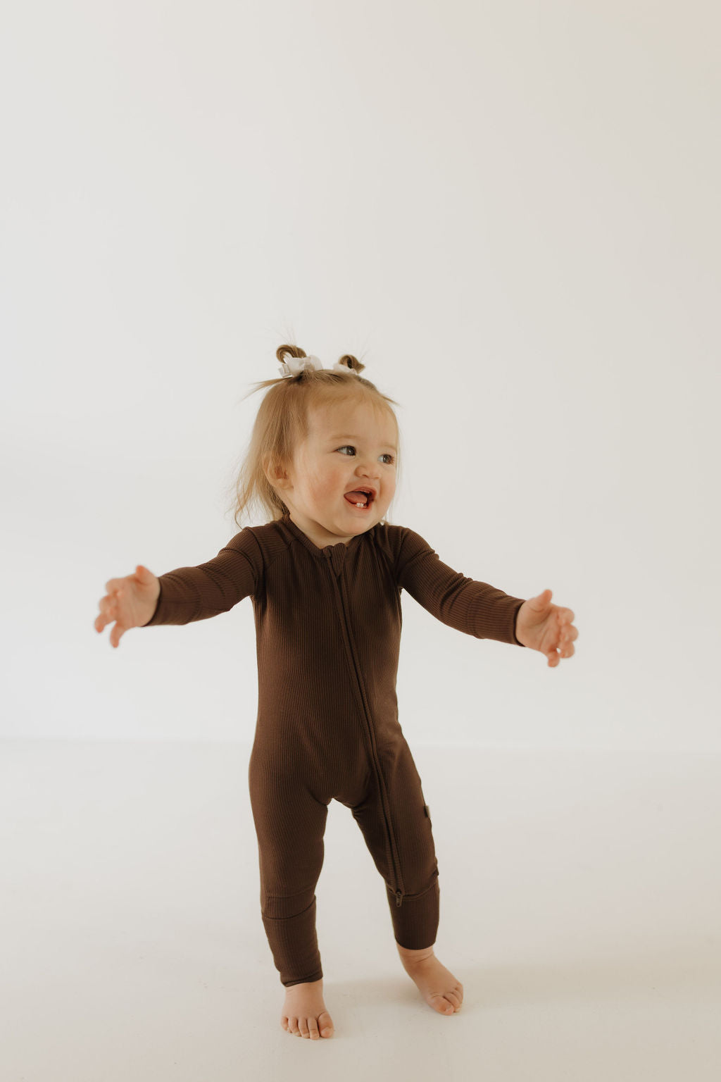 A toddler in forever french baby's Bamboo Zip Pajamas in Coffee Bean stands barefoot on a light floor with a bright background, smiling with arms outstretched and sporting two small ponytails.