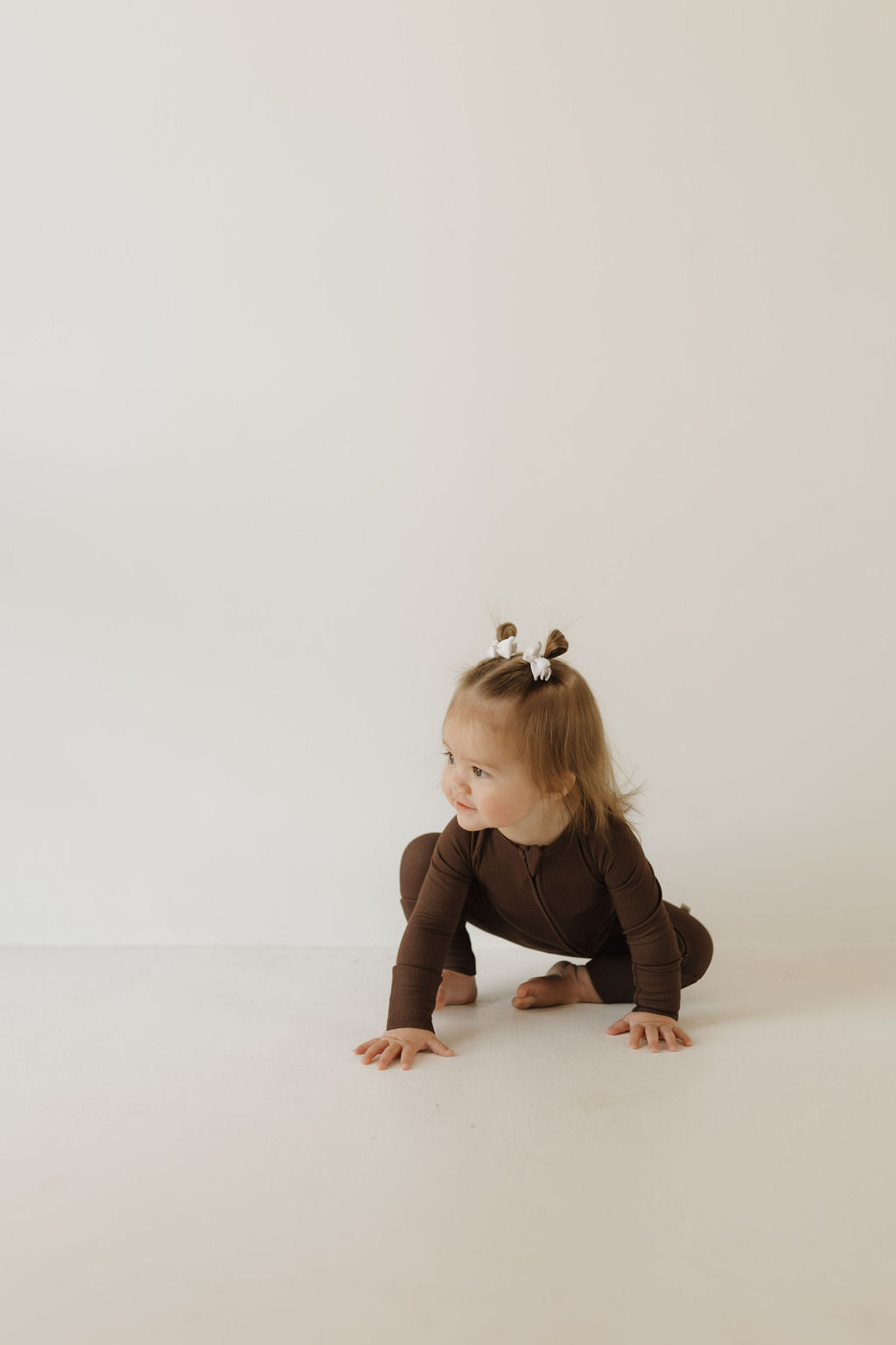 A toddler in forever french baby Bamboo Zip Pajamas | Coffee Bean is sitting on the floor, crawling. With light brown hair and small pigtails tied with white bows, the child looks curiously to the side. The hypo-allergenic fabric against an off-white background creates a soothing scene.