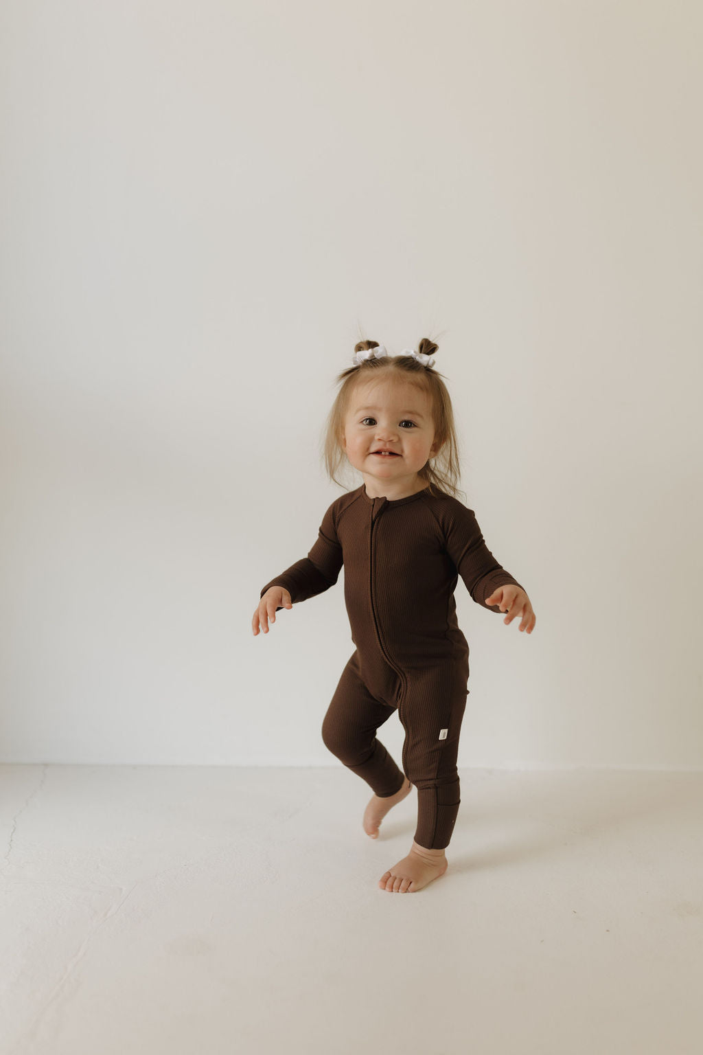 A toddler in forever french baby’s Bamboo Zip Pajamas in Coffee Bean stands on a white floor against a plain wall, smiling with hair styled into two pigtails. These hypoallergenic pajamas ensure comfort for sensitive skin.