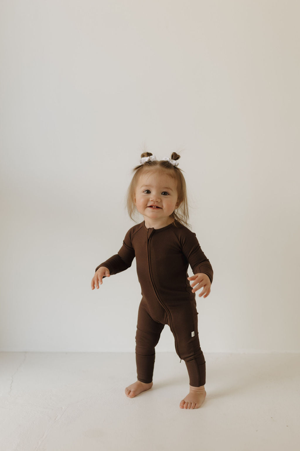 A cheerful toddler stands smiling in Forever French Baby's Bamboo Zip Pajamas in Coffee Bean. Sporting two small ponytails and barefoot on a white background, the little one embodies pure joy and comfort in this breathable, hypo-allergenic outfit.