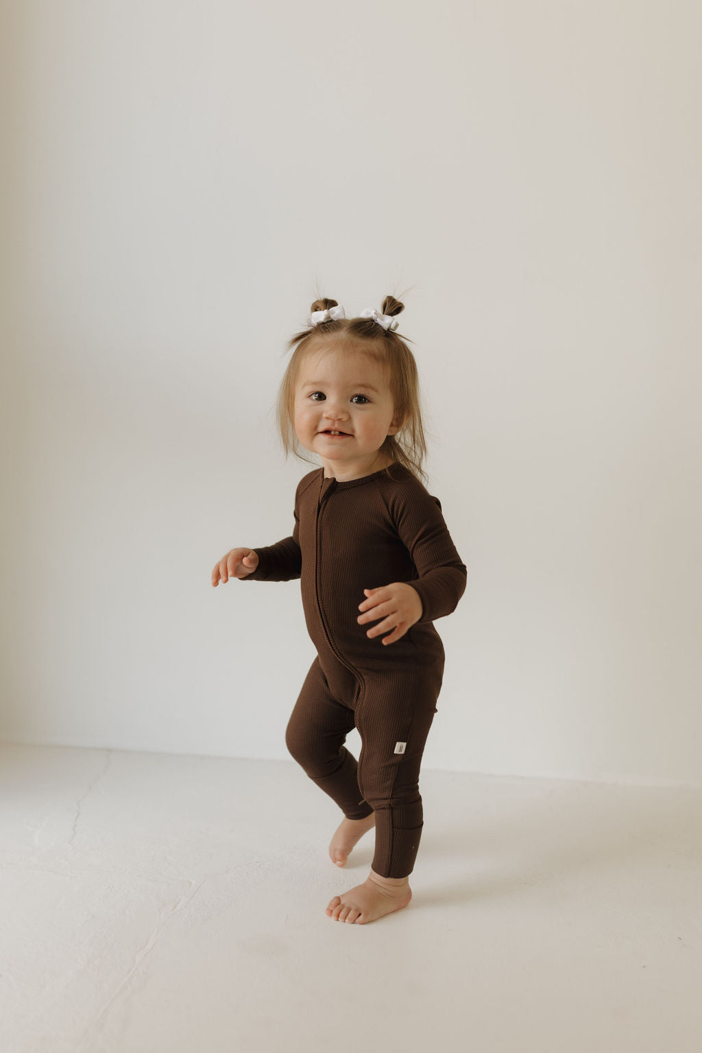 A joyful toddler in "Bamboo Zip Pajamas | Coffee Bean" by forever french baby stands barefoot on a light floor against a white background, sporting two small ponytails.