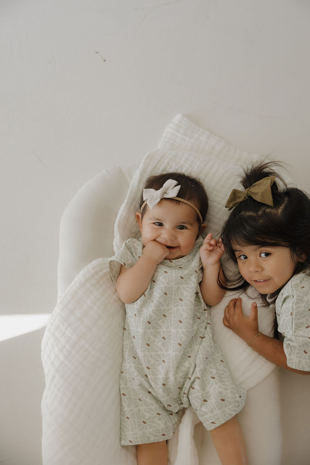 Two young children lie on a white blanket. The baby smiles in the forever french baby French Terry Romper | Coast and a white headband, next to an older child with a green bow and matching outfit—both wearing adorable newborn apparel.