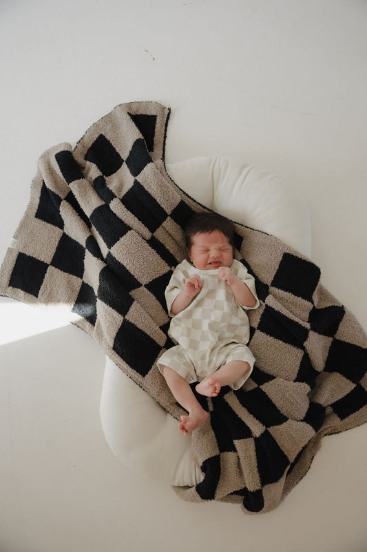 A baby sleeps peacefully on a white cushion, dressed in the forever french baby French Terry Romper | Checkerboard, featuring a light checkered print and snap closures. The scene is set against a clean white background.
