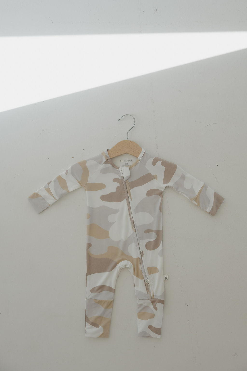 The forever french baby Bamboo Zip Pajamas | Camo 2.0 is a long-sleeve, beige and white camouflage-patterned romper with a diagonal zipper, shown on a wooden hanger against a light wall with a bright diagonal shadow.