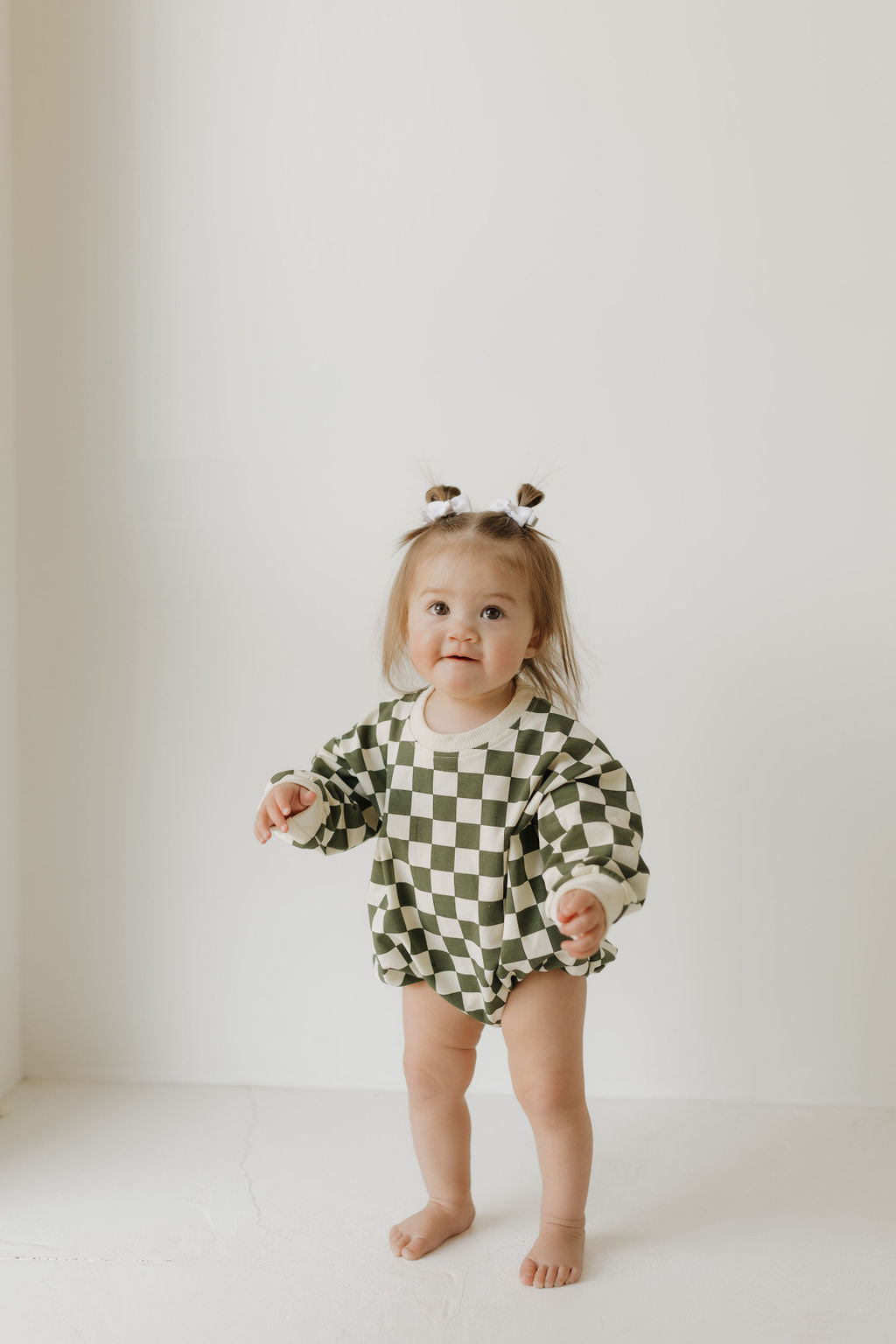 A toddler in a charming green and white checkered "Lucky" Sweatshirt Romper by forever french baby stands against a plain white background. The child, with two small ponytails and barefoot, exudes a curious and playful expression.