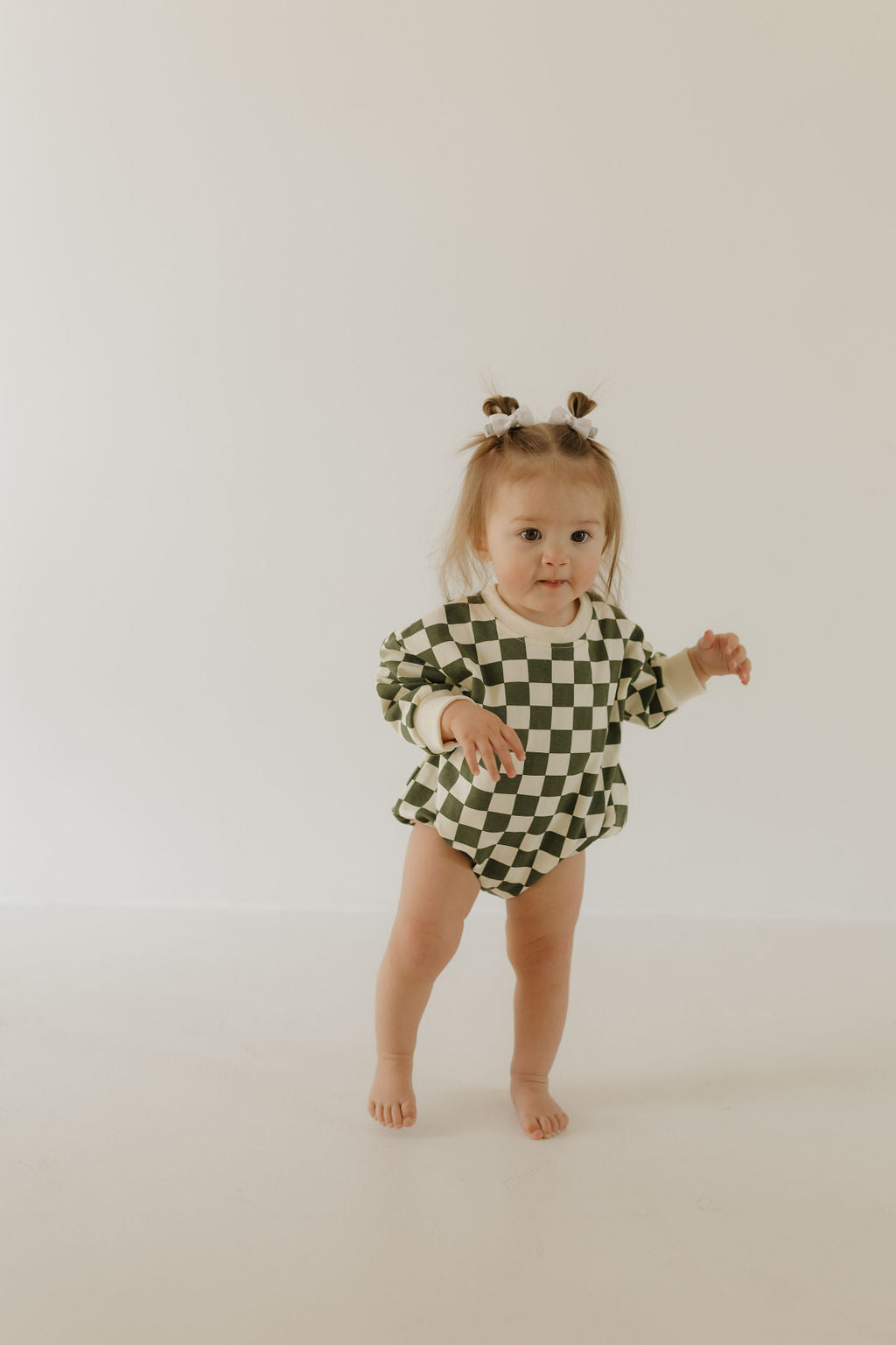 A cheerful toddler with light brown hair in two buns stands barefoot on a white surface, wearing the "Sweatshirt Romper | Lucky" by forever french baby. The green and white checkered cotton elastane outfit complements the plain off-white wall behind her.