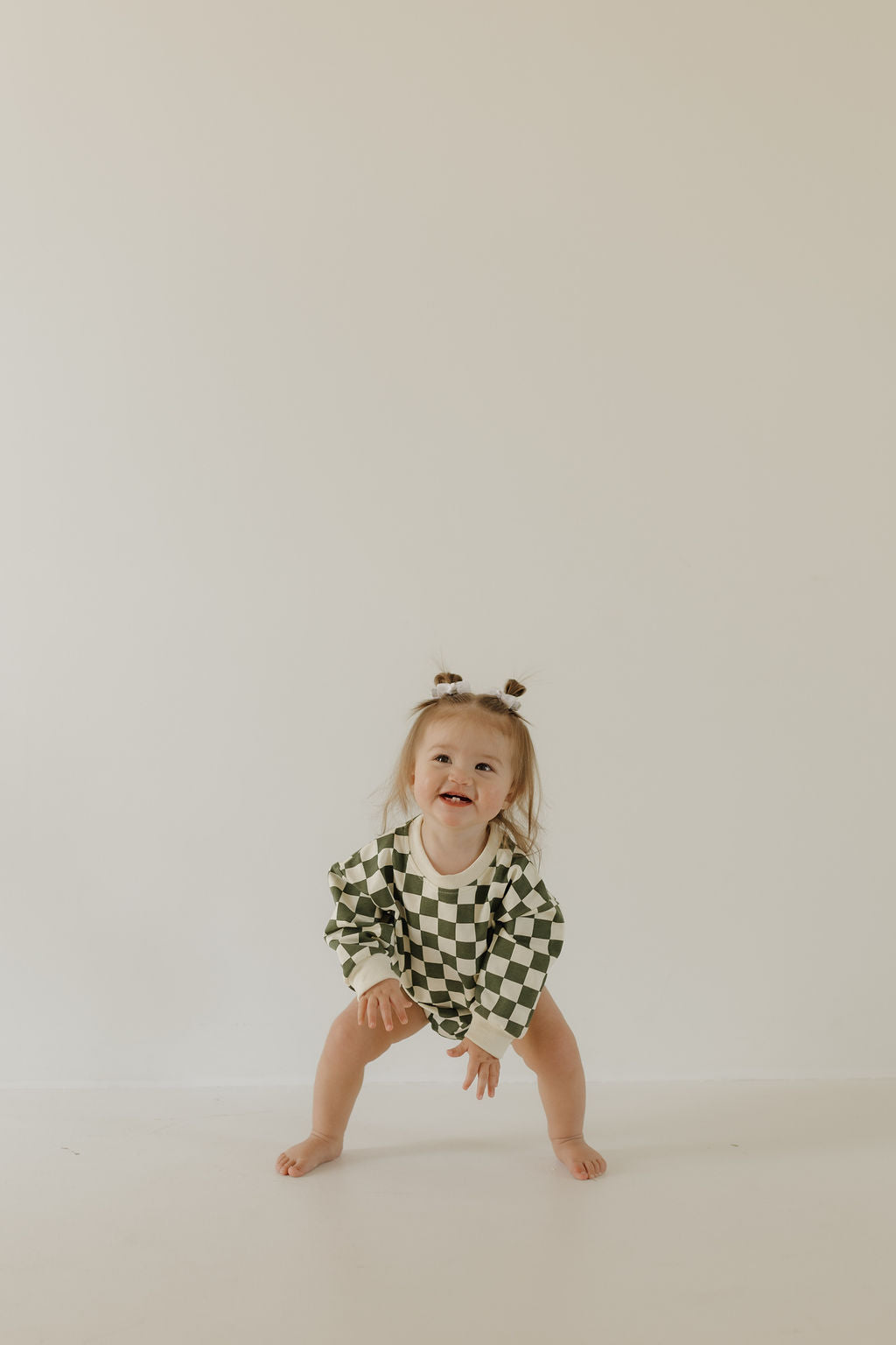 A toddler with two small pigtails poses happily against a plain backdrop, wearing the Sweatshirt Romper | Lucky by forever french baby. The green and white checkered outfit is made of soft cotton elastane, and the child stands with feet apart and hands on knees.