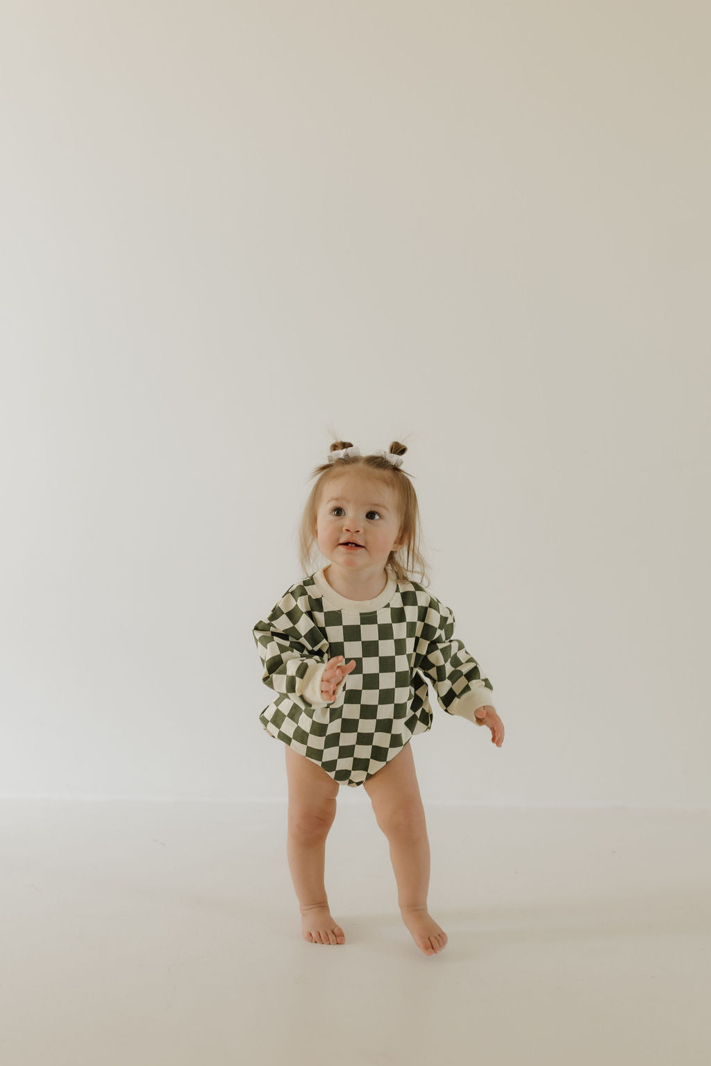 A toddler in the "Sweatshirt Romper | Lucky" by forever french baby wears a black-and-white checkered matching set on a white background, with a curious expression and two small ponytails.