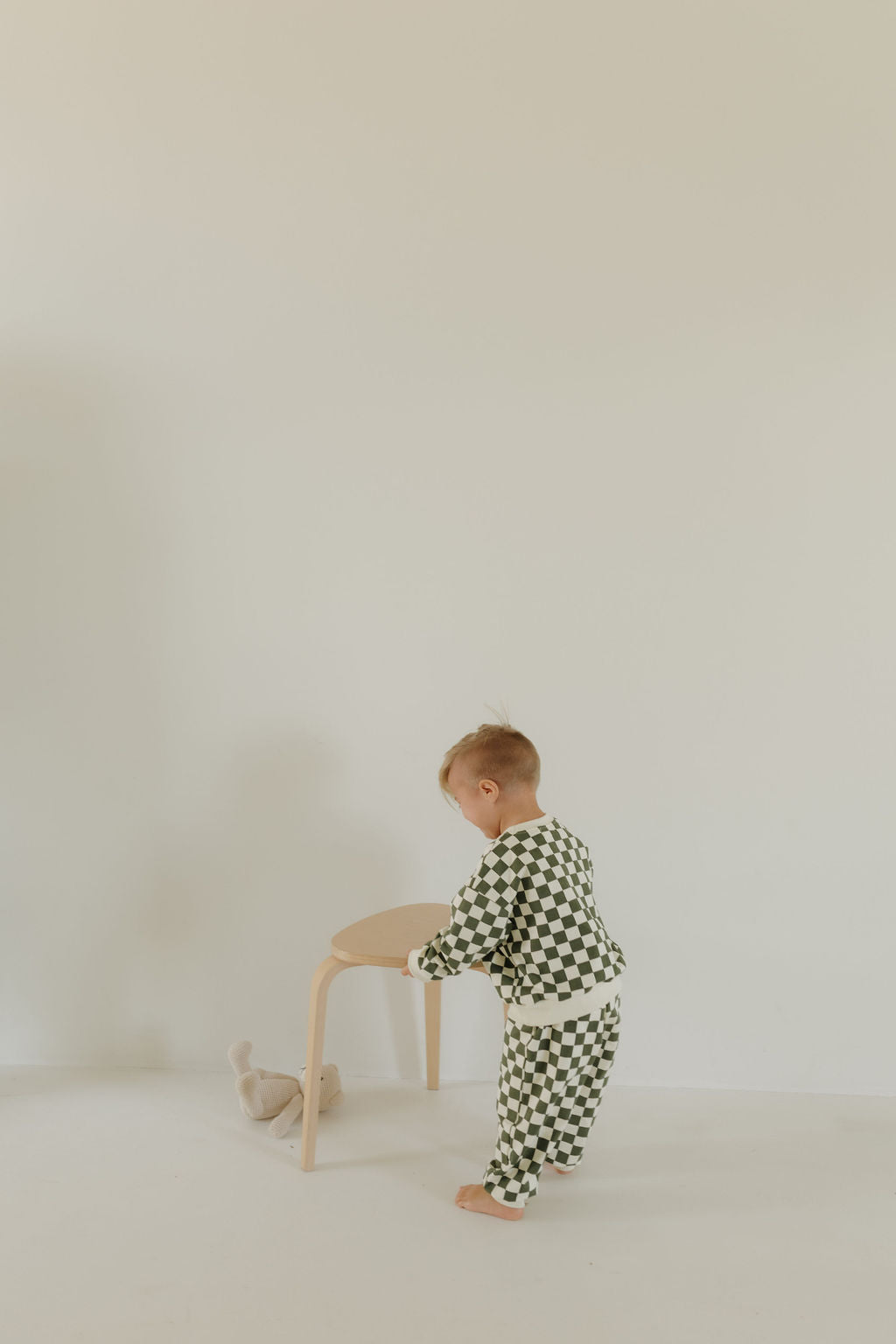 A small child in a green and white checkered Sweat Set by forever french baby stands next to a light wooden stool in a minimalistic, neutral-colored room. A soft plush toy lies on the floor beside the stool, completing this charming scene.