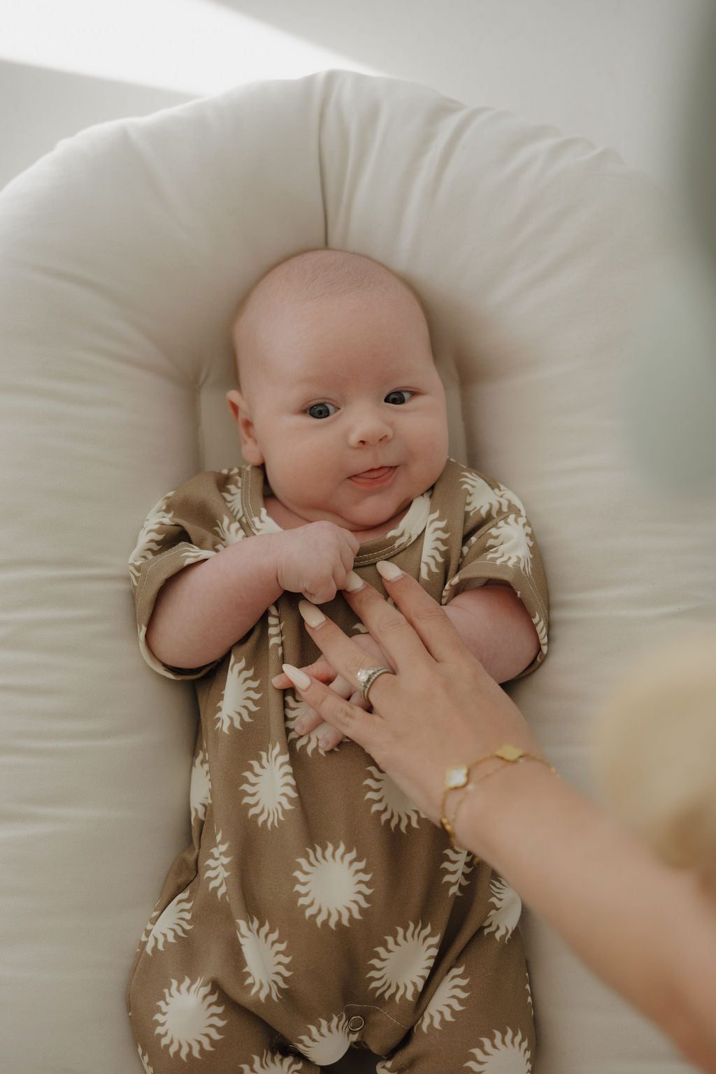 A baby in the forever french baby French Terry Romper | Sol, featuring breathable brown fabric with white sun patterns, lies on a soft white cushion while an adult gently touches the baby's chest.