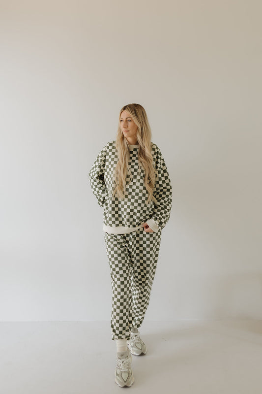 A long-haired person stands against a plain background, wearing forever french baby's Adult Sweat Set | Lucky. The checkered sweater and pants paired with patterned sneakers add a relaxed vibe, perfect for cooler weather, as they casually keep their hands in their pockets.