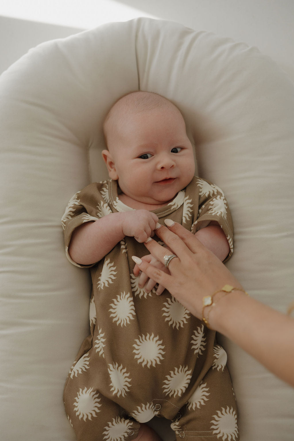 A baby in the forever french baby French Terry Romper | Sol, featuring a tan color and white sun patterns, lies on a soft white cushion, gently holding an adult's hand and gazing up with a slight smile.