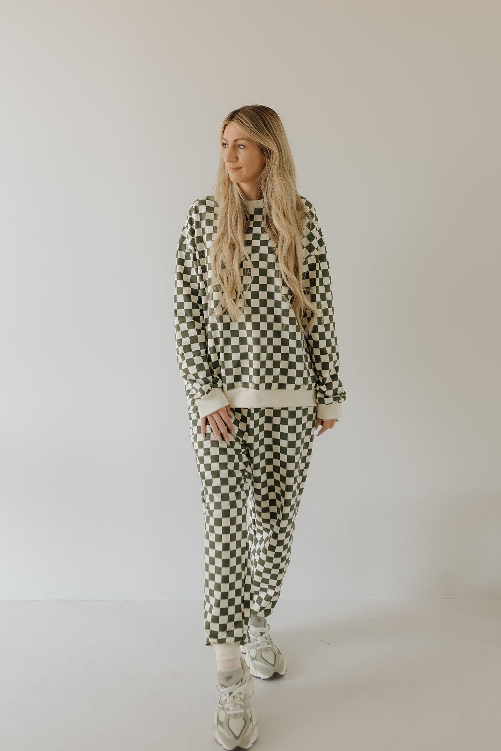 A person with long hair is against a plain background, wearing the "Adult Sweat Set | Lucky" by forever french baby, a checkered sweatshirt and pants set ideal for cooler weather, paired with sneakers.