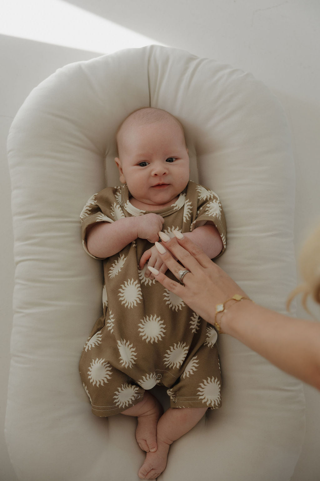 A baby wearing the forever french baby French Terry Romper | Sol, featuring a tan shade with a white sun pattern, lies on a soft surface as an adult hand with a ring and bracelet gently rests on the baby's chest.