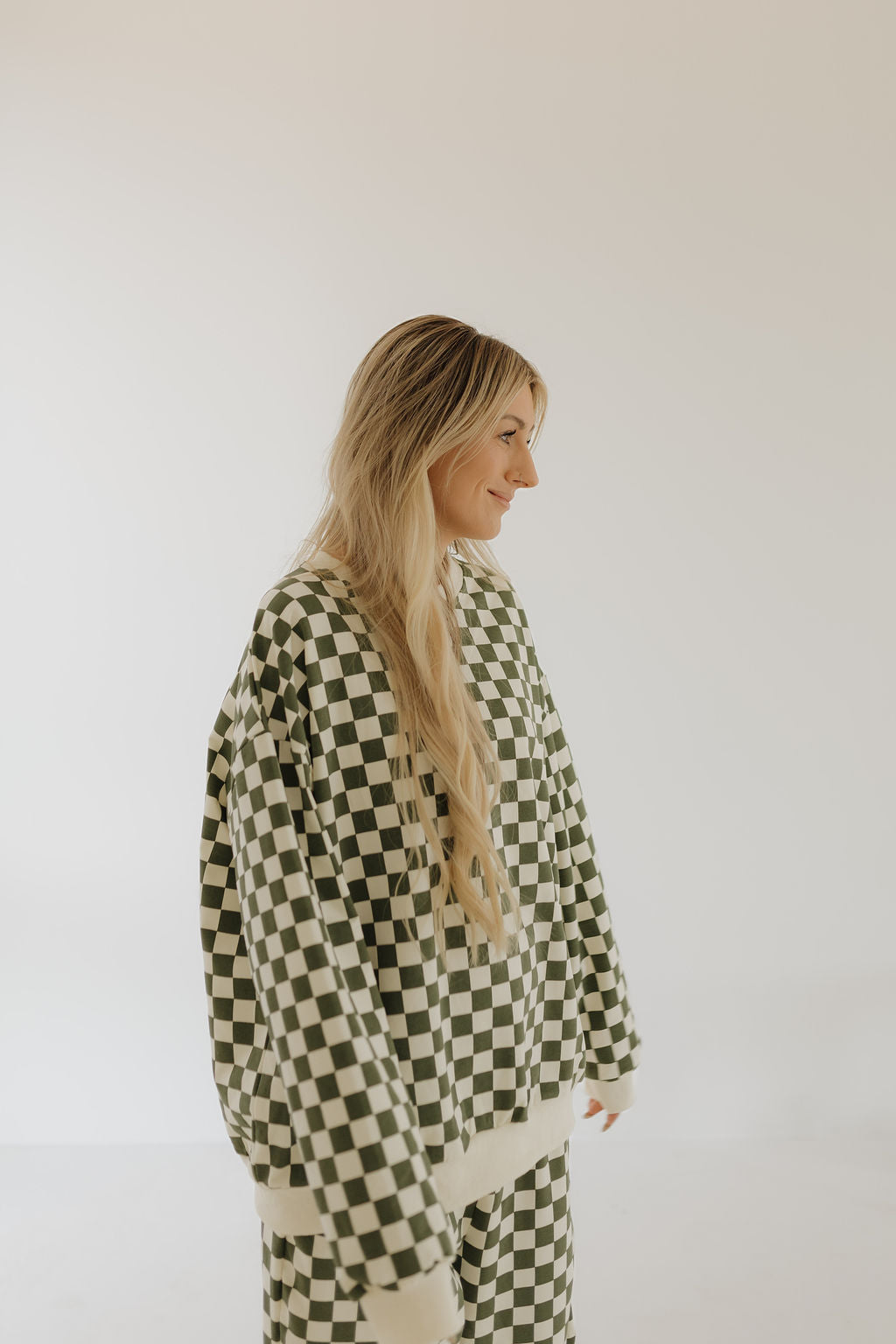 A person with long blonde hair wears the “Adult Sweat Set | Lucky” by forever french baby, featuring a matching black and white checkered design ideal for cooler weather. They stand against a plain light background.