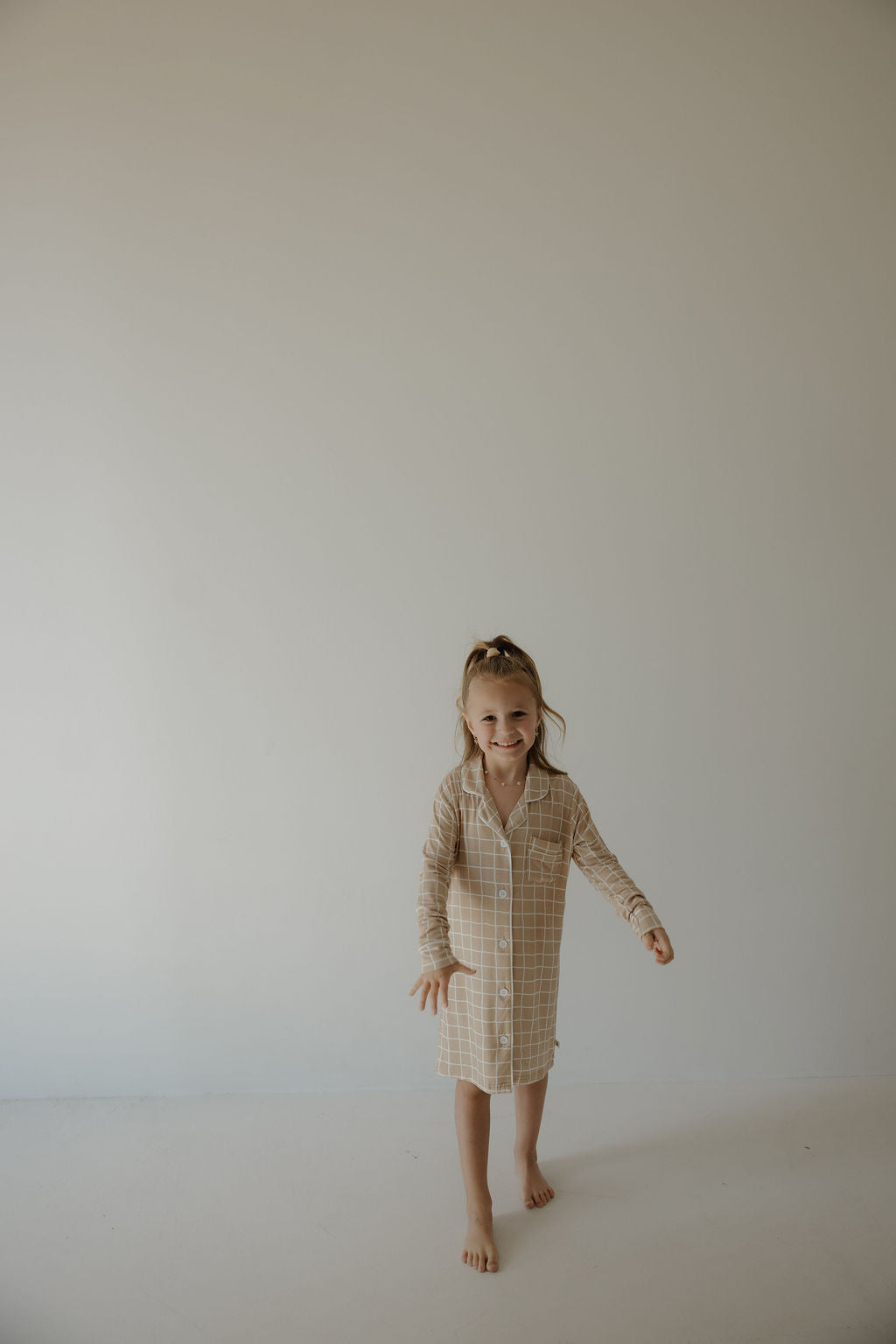 A young girl with light brown hair in a ponytail smiles barefoot in the forever french baby "Toddler Bamboo Sleeping Dress | Grid," a hypo-allergenic beige plaid nightshirt, against a plain, light-colored background.